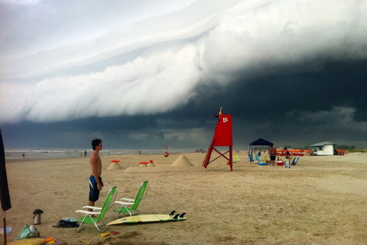 A sandy beach under a huge, low wall of dark storm cloud rolling in from the sea. A shirtless young man in blue shorts stands looking toward the water next to two green folding chairs and a surfboard lying on the sand. A red lifeguard tower stands behind him, and a few people gather near a small blue tent further down the beach.