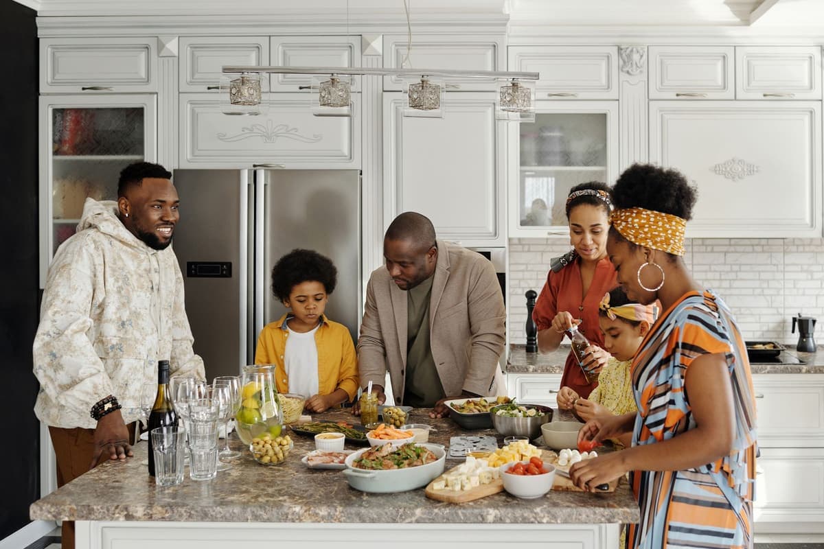 A family of six gathers around a kitchen island covered with dishes of salad, cheese cubes, cherry tomatoes and olives. A smiling man in a light hooded jacket stands on the left, a boy in a yellow shirt watches a man in a beige blazer lean over the food, and on the right a woman opens a bottle while another woman in a striped dress and patterned headscarf prepares tomatoes next to a young girl.