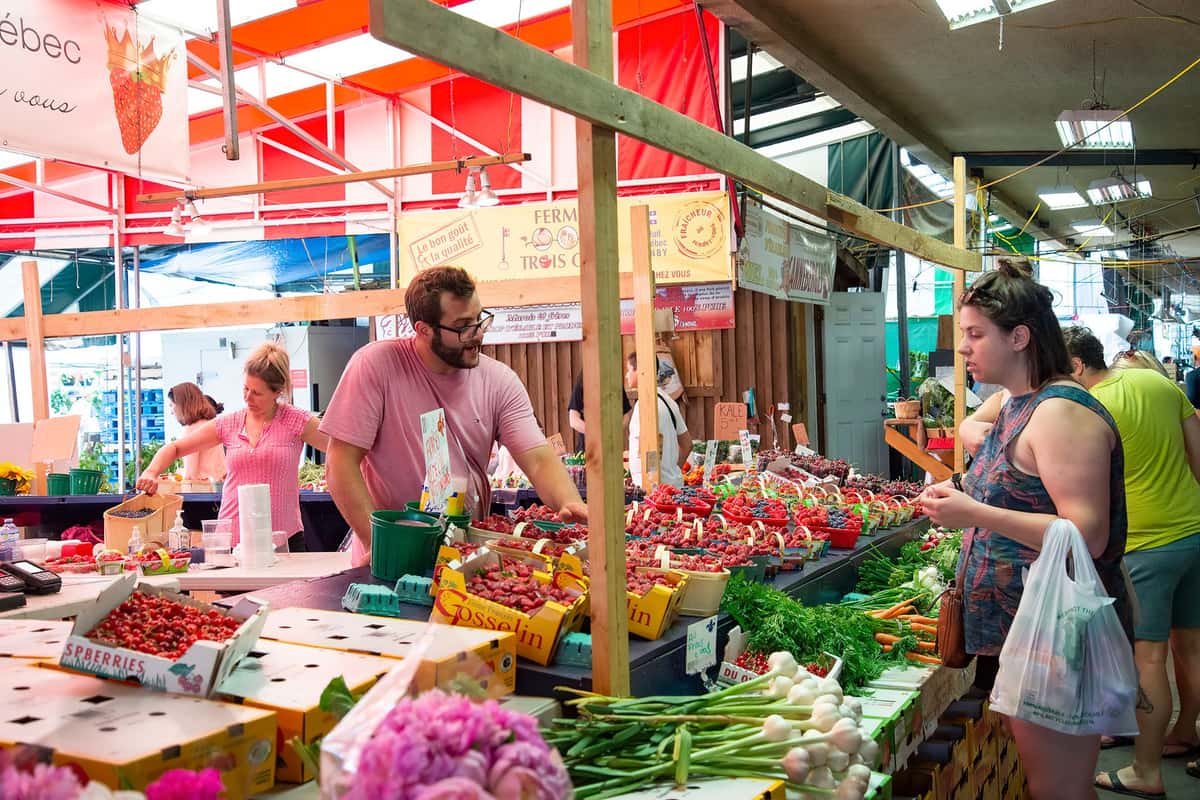 A busy covered farmers' market stall. A woman in a pink top arranges fruit at the back while a bearded man in a pink T-shirt and glasses reaches across trays of strawberries and raspberries. On the right, a woman holding a plastic shopping bag looks at bunches of carrots, green onions and herbs as other shoppers pass behind her.