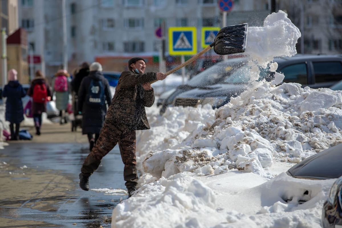 On a city sidewalk after a snowstorm, a man in a camouflage jacket and cap swings a large shovel, throwing a heap of snow into the air toward a snowbank that half buries a row of parked cars. Behind him, several people in winter coats, some with backpacks, walk along the wet sidewalk past a pedestrian-crossing sign.