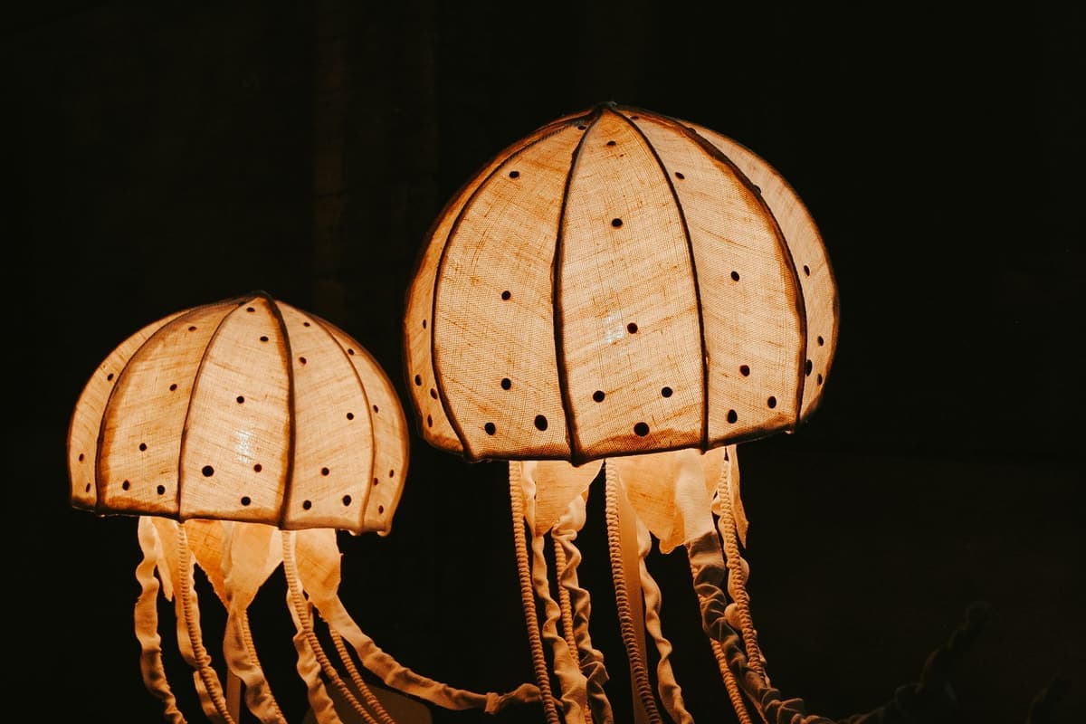 Two glowing lamps shaped like jellyfish against a black background. Each has a round, dome-shaped shade made of pale woven material with rows of small holes, lit from inside with a warm orange light. Long, wavy strips of fabric hang down below each shade like tentacles.