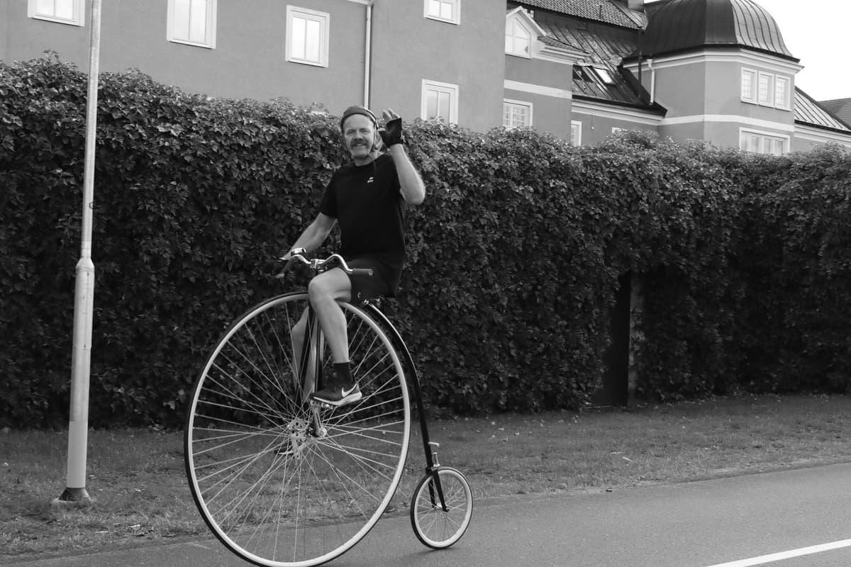 A black-and-white photo of a smiling man with a moustache riding a penny-farthing bicycle along a street and waving. The front wheel is enormous, about as tall as his chest, with the seat on top and the pedals fixed to its centre, while the back wheel is tiny. A tall hedge and a large building are behind him.