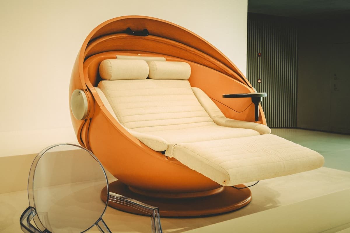 A futuristic reclining lounge chair in a showroom. The chair is a large glossy orange shell shaped like an open egg, set on a round wooden base, with a cream padded seat and two cushions that stretch out into a long footrest. Round speakers are built into both sides of the shell, and a small black tray sticks out on the right.