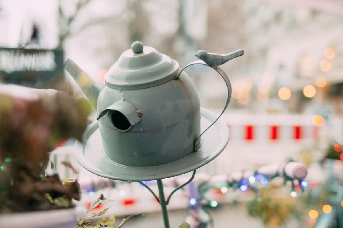 A pale green metal birdhouse shaped like a teapot, standing on a saucer at the top of a thin metal pole. There is a round entrance hole on the front, a lid with a knob on top, and a small metal bird sits on the curved handle. Behind it, coloured lights and a red-and-white barrier are blurred.