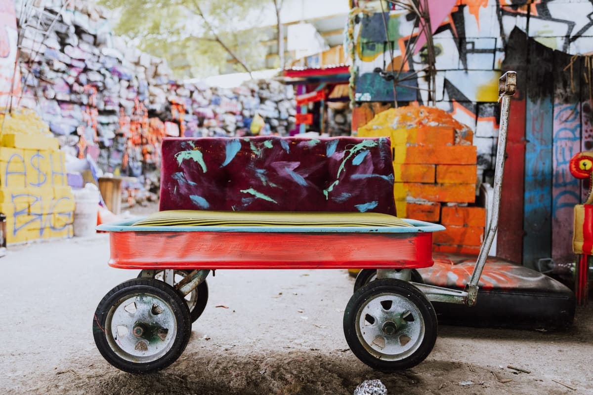 A red metal children's wagon turned into a bench, parked in a concrete courtyard. It has four small rubber wheels, a long metal pull handle on the right, a flat yellow-green seat cushion, and a tall dark red backrest painted with green and blue leaves. The walls behind it are covered in colourful graffiti.